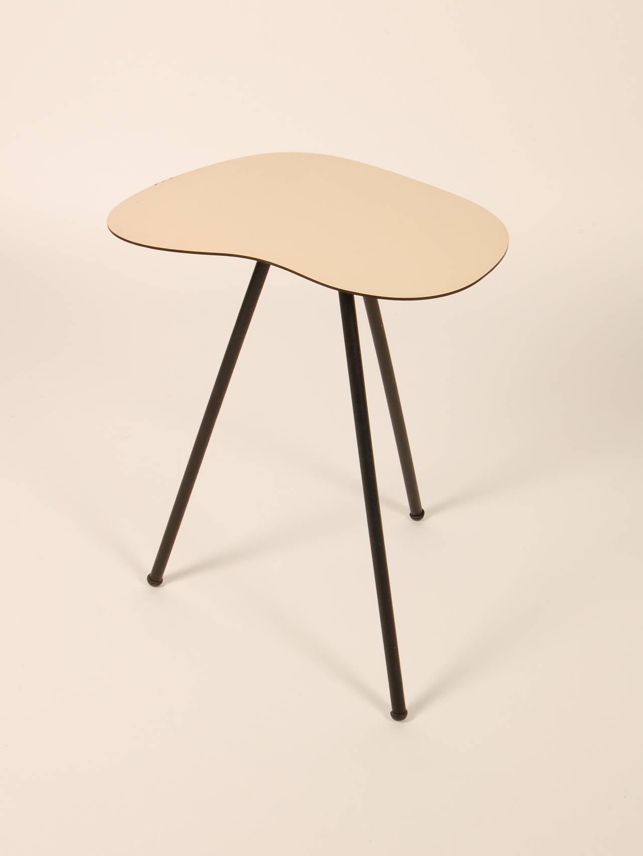 Petite three legged side table with a free form top. Constructed of an off white laminate top and black lacquered wood legs. Wonderful painters palate form with a very 1950s approach to the three legged table. Labeled by the marker.