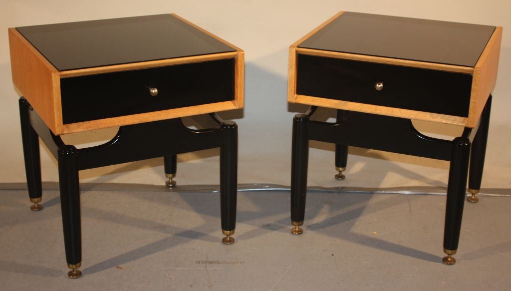 A chic pair of ebonized and natural wood side stands with black mirrored tops and brass details, each with a single drawer, each stamped E. Gomme, Wycombe.