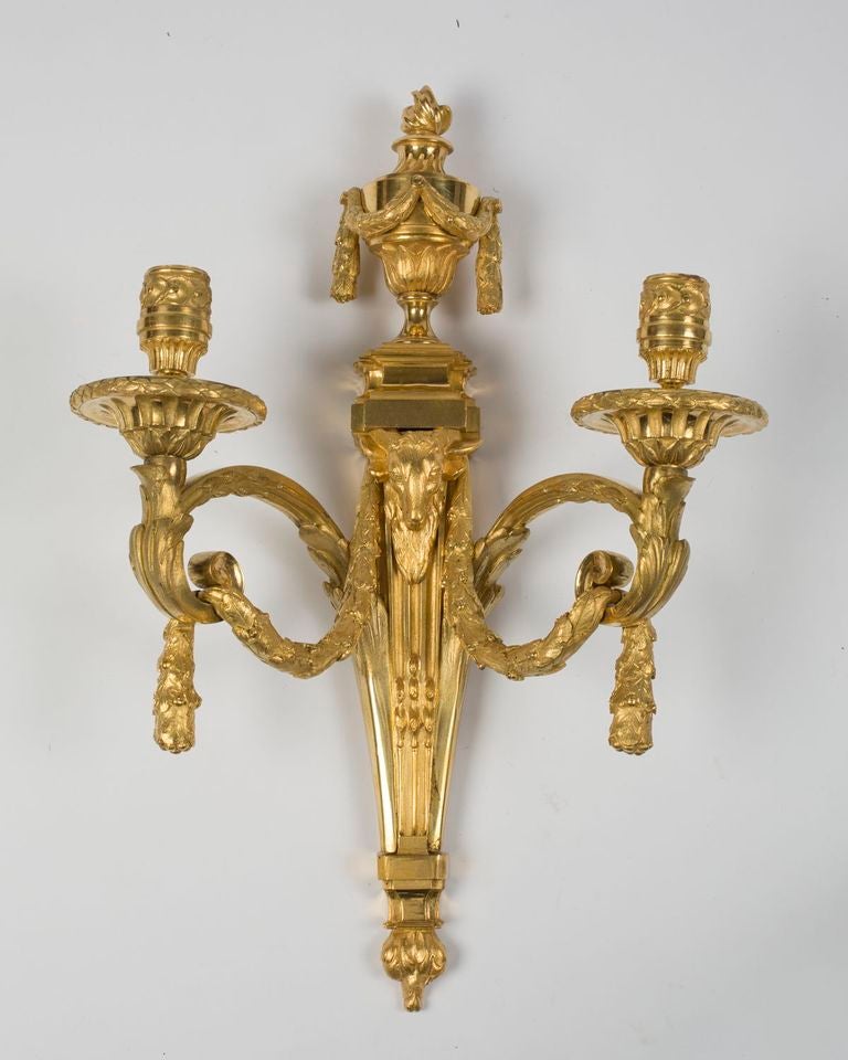 Each surmounted by a flaming classical urn hung with laurel swags over the tapering fluted backplate cast with the head of a ram, issuing scrolling foliate candle arms joined by laurel-leaf garland, ending in leaf-cast drip-pans.  After a design by