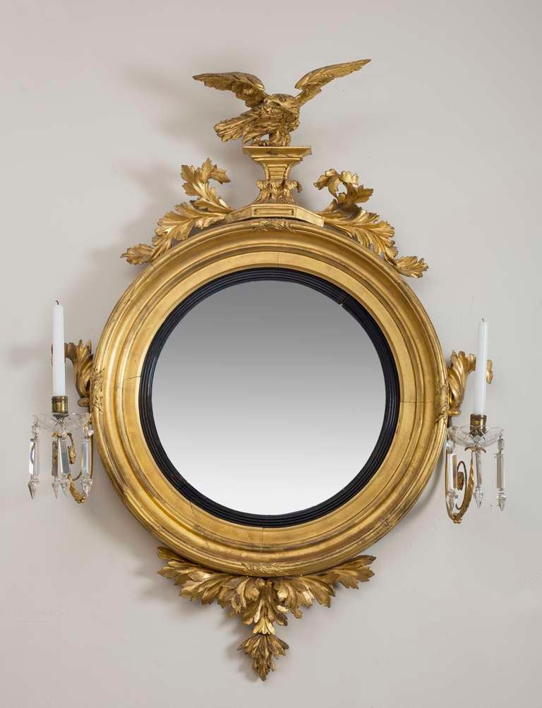 With a fully sculpted eagle perched above foliate scrolls, the circular molded mirror frame surrounding a convex mirror plate, the frame affixed with brass candlearms hung with glass prisms above a scrolling foliate apron. 