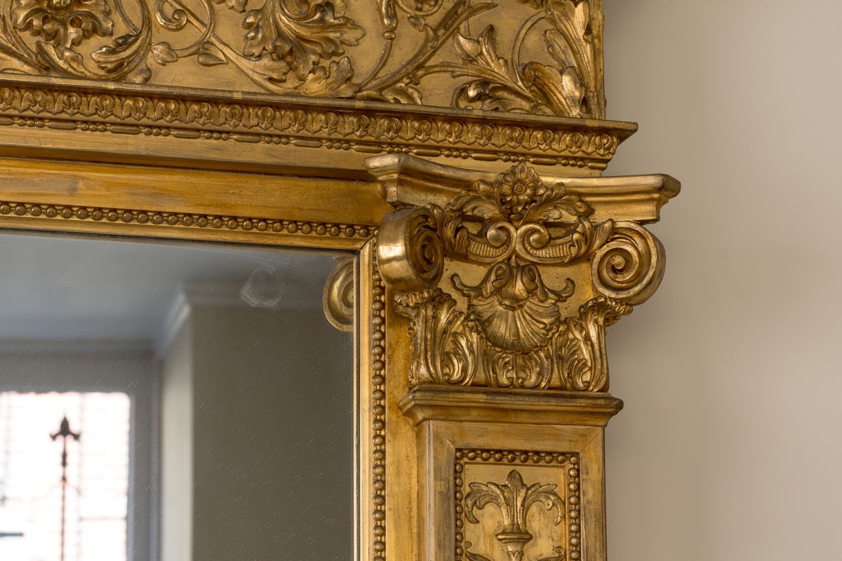 The rectangular modern mirror plate surmounted by an egg-and-dart pediment above a foliate and floral arabesque frieze, flanked by two pilasters carved with reeded urns with scrolling branches centered by a mask and ending in Corinthian capitals,