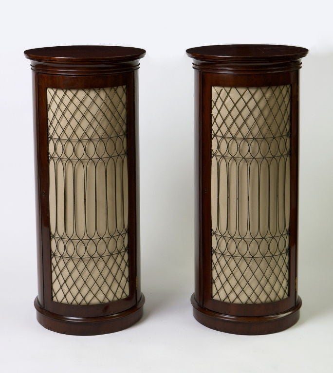 Each of cylindrical shape, the circular top above a single brass grille door lined in beige silk opening to reveal two shelves, raised on a plinth base.