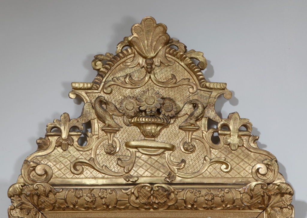 The later rectangular mirror plate within a C-scroll and diaper pattern carved border surmounted by a cresting carved en suite and centered by a vase with three sunflowers, topped by a stylized scallop.