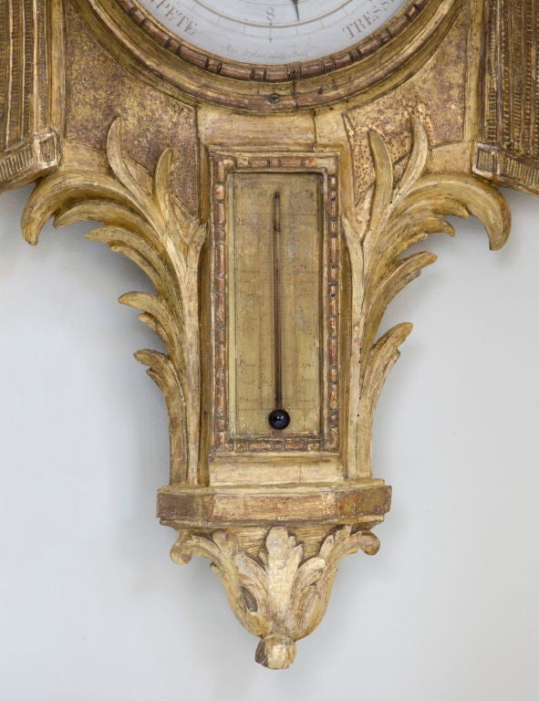 Of typical form‚ the giltwood body centered by a circular barometer dial flanked by pillars ending in flame‚ above a thermometer flanked by foliate branches‚ surmounted by swags.