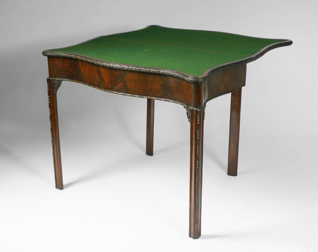 The serpentine rectangular hinged top with a flower-carved edge enclosing a green baize-lined playing surface, on square canted legs with foliate carved corner brackets.