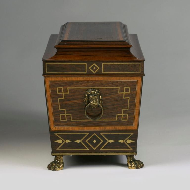 Of sarcophagus form with geometric brass stringing throughout, with embossed gilt-brass ring handles to the sides, the interior fitted with twin lidded lead-lined caddies separated by a glass mixing bowl, raised on brass paw feet. With a secret