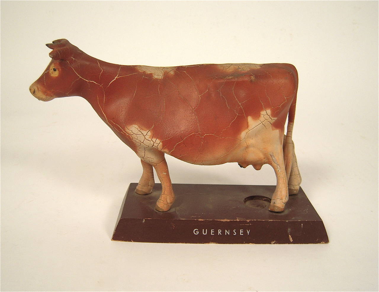 A German model (or toy) of a Guernsey cow, well modeled in painted plaster or cardboard (?), with an old, crackled leathery surface, on a painted wood plinth with 'Guernsey' stenciled in silver letters on it. 'Made in Germany' stamped on base. There