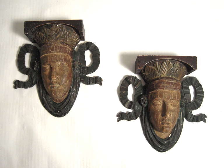 A pair of well carved and painted neoclassical mask wall brackets or wall lights, likely from a theater, each idealized face surmounted by carved stylized acanthus leaves, flanked by carved tied ribbon and over a carved and painted linenfold swag.