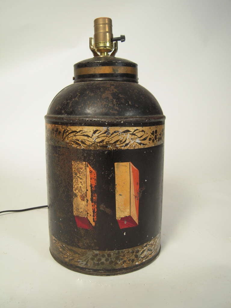 A 19th century English 'tôle peinte' tea canister, fitted as a table lamp, the cylindrical form with a domed cover, decorated with the shaded number '11'in  gilt  and red, with decorative gold foliate bands on a black painted ground.