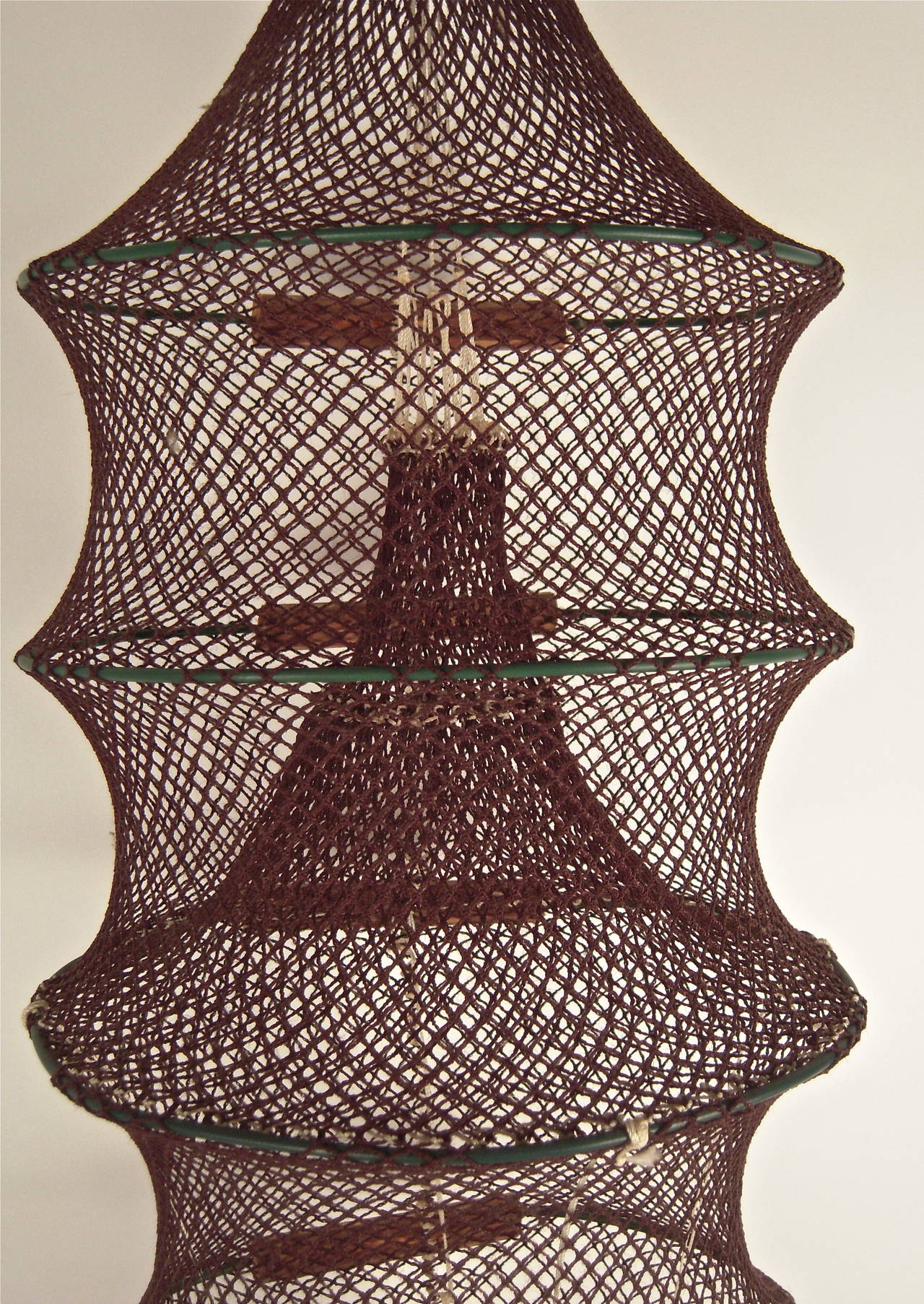 A sculptural, handmade, knotted string fish trap, the conical, cylindrical form in brown and red hand knotted string on circular green metal frames, with cylindrical wood back supports, the bottom weighted with a semicircular wood frame and red