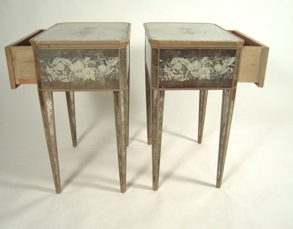 A stylish pair of  1940s end or night tables, of rectangular form with canted corners, raise don square section tapering legs, each with mirrored surface overall, decorated with reverse glass white stenciled floral and foliate decoration, the tops