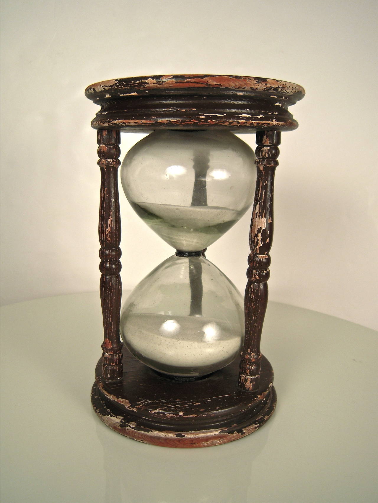 A large 19th century hour glass, or sand glass, with brown painted ring-turned end plates connected by three baluster- turned columns, encasing a large colorless glass bulb with white sand.

Height: 12 3/4