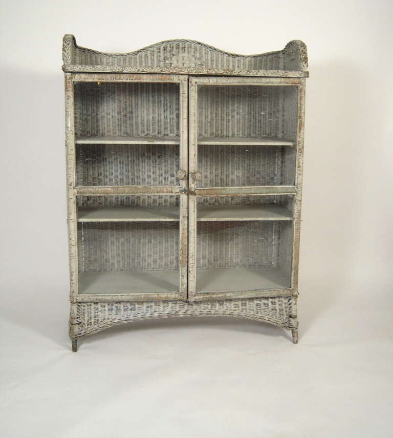 A 19th Century American painted wicker cabinet, in old white paint, retaining its original, wavy glass doors, the serpentine cresting and sides over a 3 shelf cupboard with double doors, supported by a gently arched base raised on 4 legs.