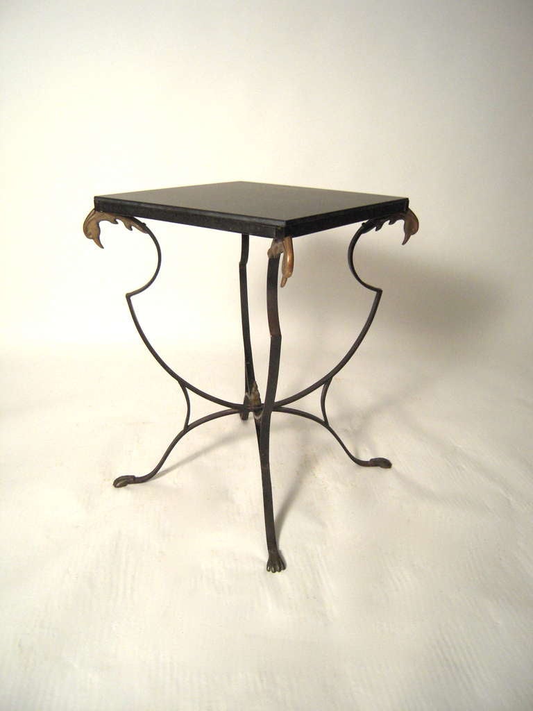 A stone-top side table attributed to Maison Jansen, the square top with later replaced soap stone supported by a graceful black strap iron base with brass swan's heads, the serpentine supports joined by a stretchers centered by a brass flame finial,