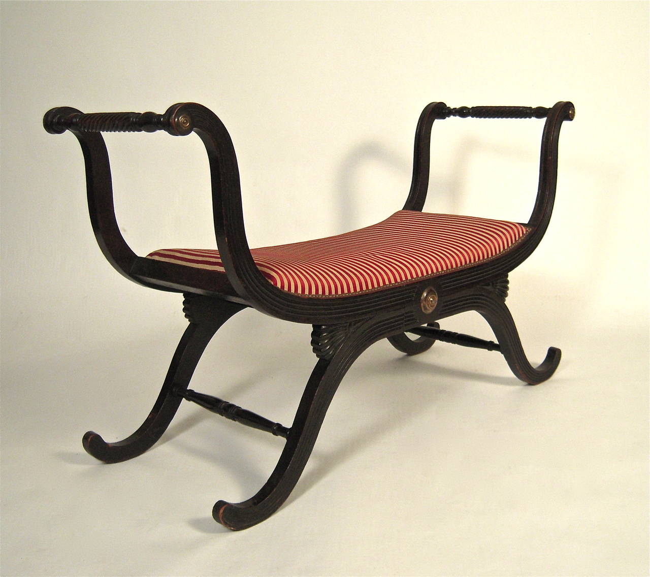 A well made and beautifully proportioned neoclassical style dark stained, carved wood window seat or bench of curule form, the reeded, elegantly curved arms, joined by spirally turned, carved supports, the upholstered seat over a brass boss on each