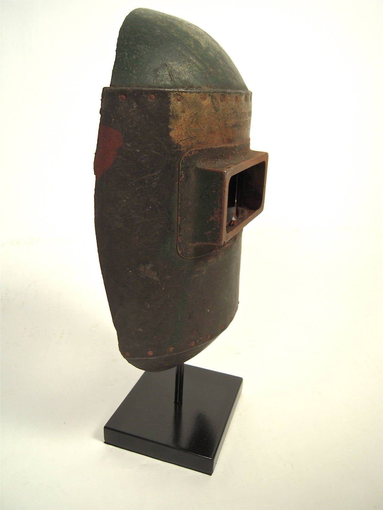 A sculptural, Industrial welder's mask on stand, American, circa 1920s, in olive green painted fiber board and metal with the manufacturer's name in block letters above the eye opening: "The Fiber Metal Products Company, Chester, PA".