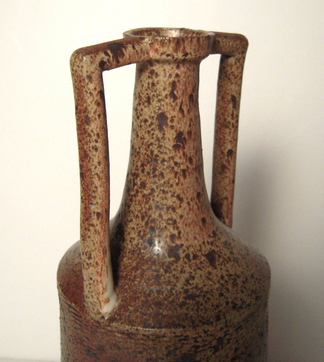 A tall neoclassical style stoneware amphora, circa 1960s-1970s with mottled brown and tan glaze and incised semicircular decoration, the bottle opening above an everted neck flanked by geometric handles above a cylindrical body tapering down to a
