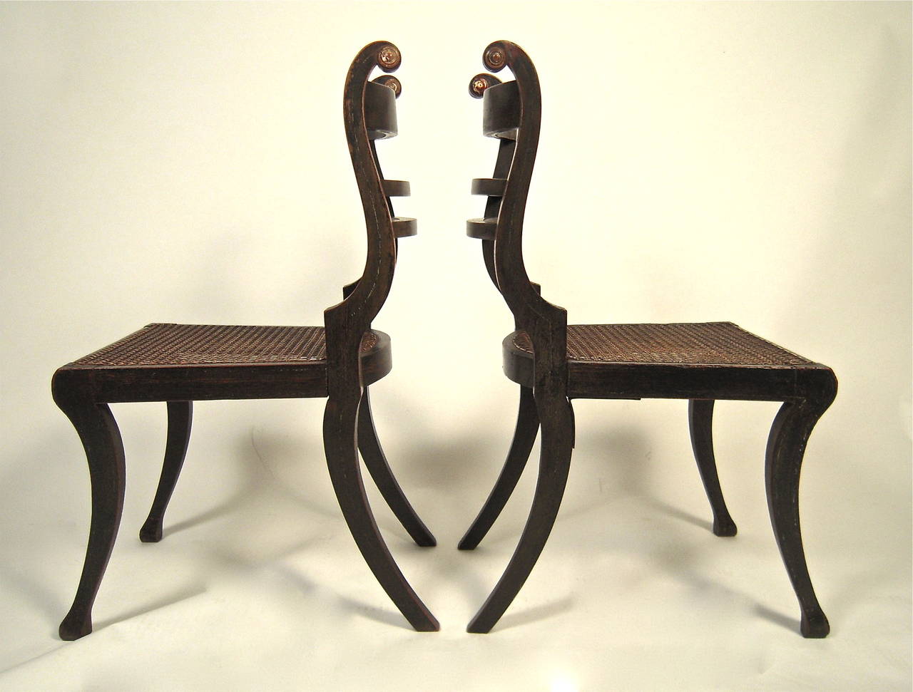 A pair of wonderfully sculptural English Regency period Trafalgar chairs, painted black with grain painted panels and traces of gilded and white pinstripe decoration, the exaggerated scrolled backs with curved slats over caned seats supported by