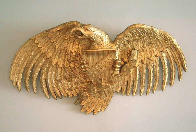 A large and beautifully carved and gilded wood American Eagle plaque, with wings spread wide, head cocked to the right, clutching the American stars and stripes shield, lightning bolts and a laurel garland in its talons.

Length: 39