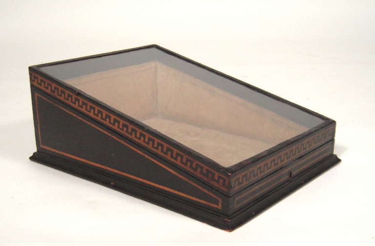 An English Regency period painted and ebonized wood tabletop display box, the glass angled cover opening to a velvet-lined interior, the ebonized case decorated with a Greek key/fret border, the sides with conforming borders. With its original key