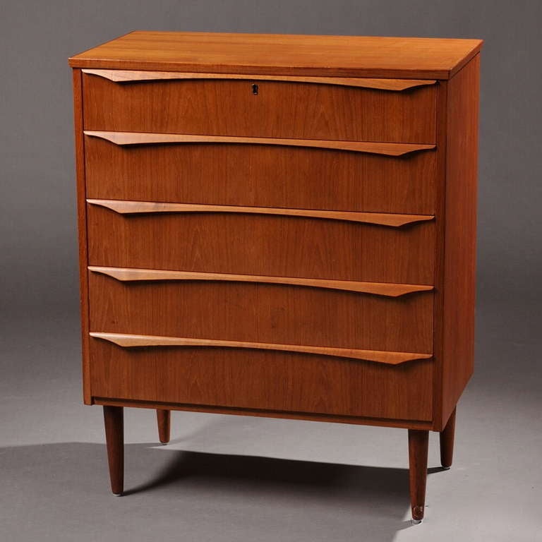 A sculptural and well proportioned Danish midcentury modern chest of drawers in  teak, the case fitted with five drawers having projecting shaped pulls along the upper edges, raised on turned tapering legs.
