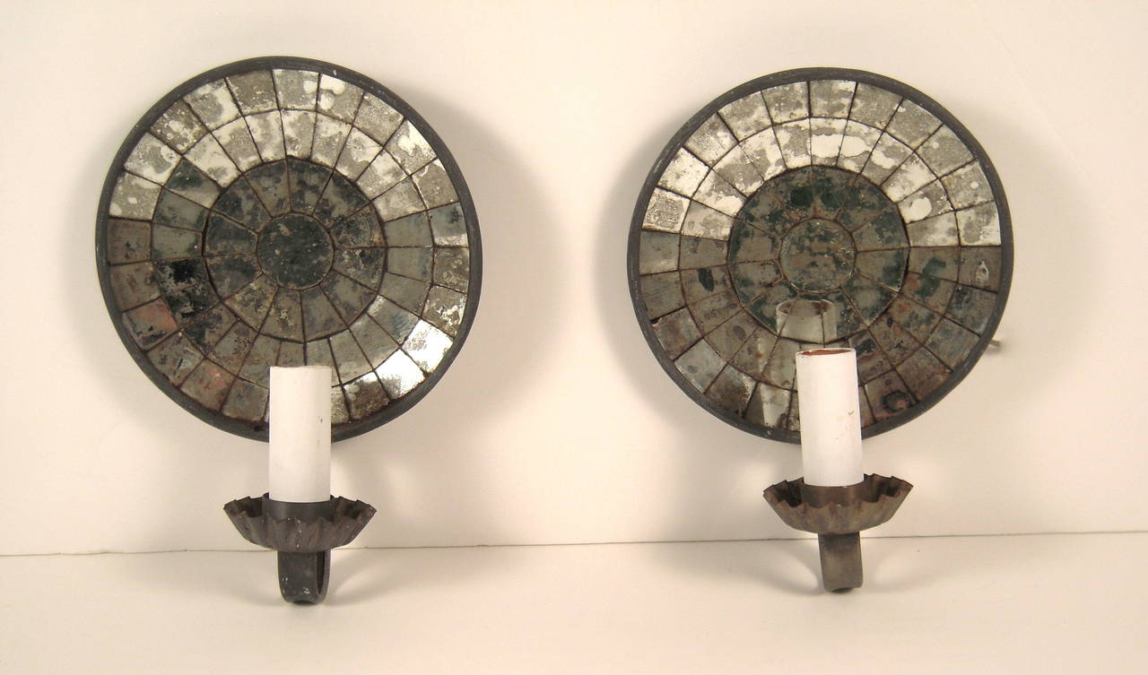 A pair of round mirror backed and tin circular wall sconces, American, circa 1930s, with white painted wood candles arms supported by tin bobeches and arms. Electrified. These sconces would be equally at home in a traditional or in an eclectic