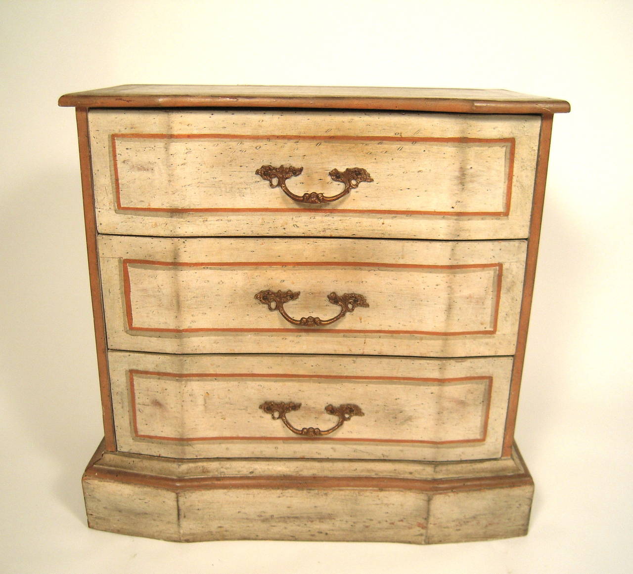 A diminutive painted Italian chest of drawers, in pressed wood, circa 1940s, the top, sides and angled three front drawers all painted in cream with terra cotta and grey and 'antiqued.' Brass drawer pulls. Perfect as a side table or nightstand,