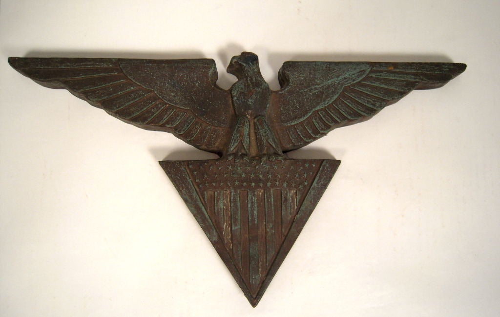 A 1930s patinated cast bronze architectural ornament in the form of a stylized American bald eagle with outstretched wings surmounting a triangular shield decoraated with the stars and stripes.