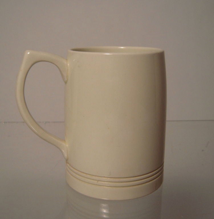 A KEITH MURRAY DESIGNED QUEENSWARE MUG FOR WEDGWOOD, THE FLARED CYLINDRICAL BODY WITH THREE INCISED LINES AT THE BASE AND A SHAPED STRAP HANDLE. Signed on base with printed mark and retaining Earl Buckman collector’s inventory decal. English, circa