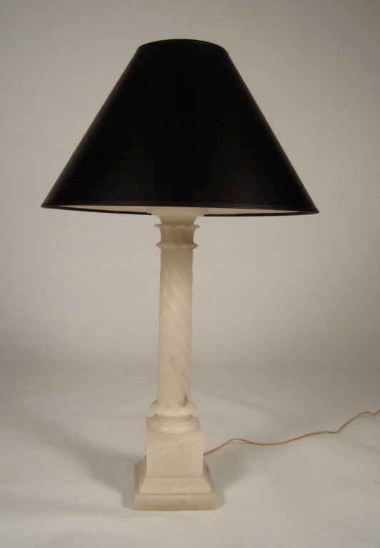 A tall carved alabaster Corinthian column lamp, the stylized carved acanthus leaf capital  over a spirally turned shaft supported on a raised square section plinth, with a stone finial and black paper shade.