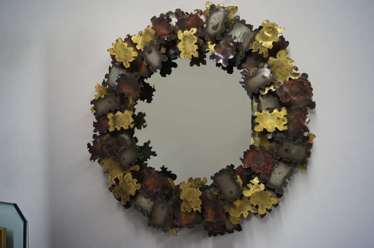 Reminiscent of the Brutalist work of Silas Seandel and C. Jeré, this circular wall mirror comprises a wreath of jigsaw-shaped patinated metal pieces in an array of tones. 

The approximate interior diameter of the mirror surface is 13 inches.