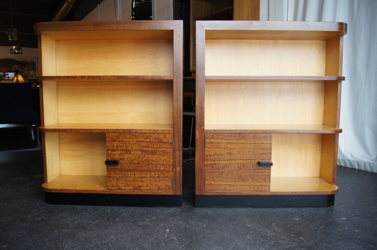 Designed by Gilbert Rohde for Herman Miller in the early 1940s, this handsome pair of art deco bookcases features a bookend design, each case with one curved corner and one lower cabinet. The exteriors of the bookcases are a dark walnut, the