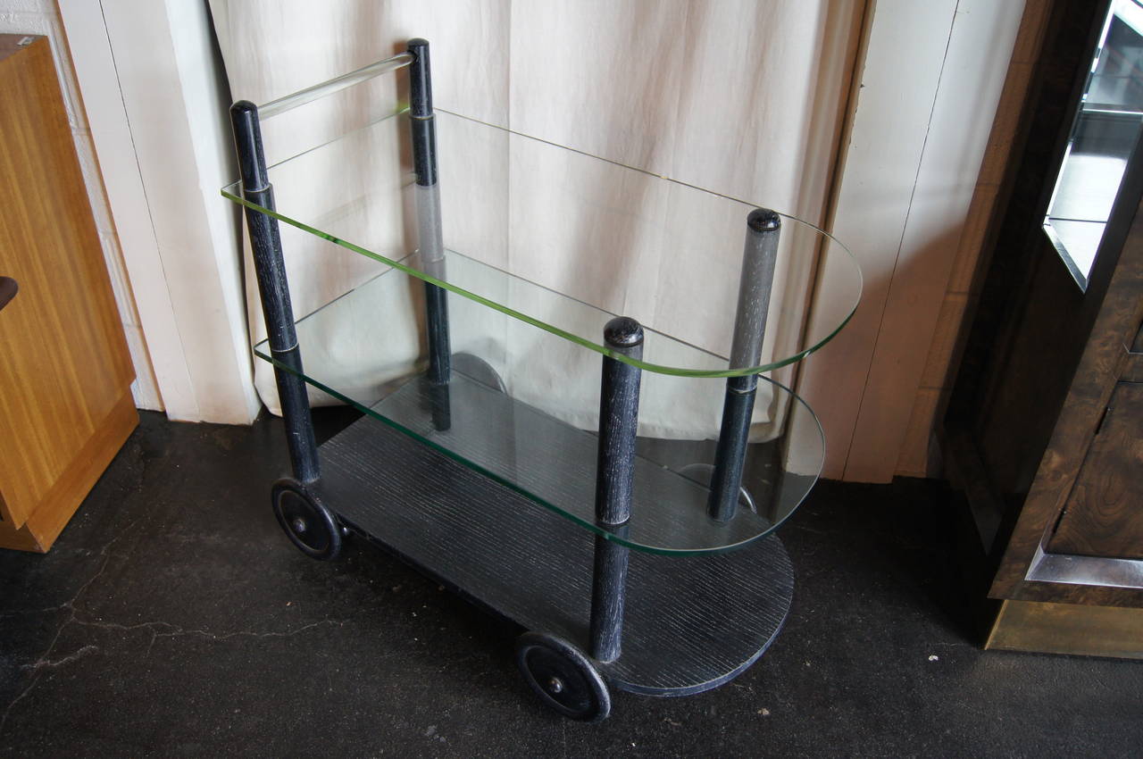 In the style of Gilbert Rohde in the 1930s, this tea cart comprises an ebonized oak frame, two glass shelves, and a glass handle. The cart features large wood wheels that conceal small casters, which were added at a later date to preserve the