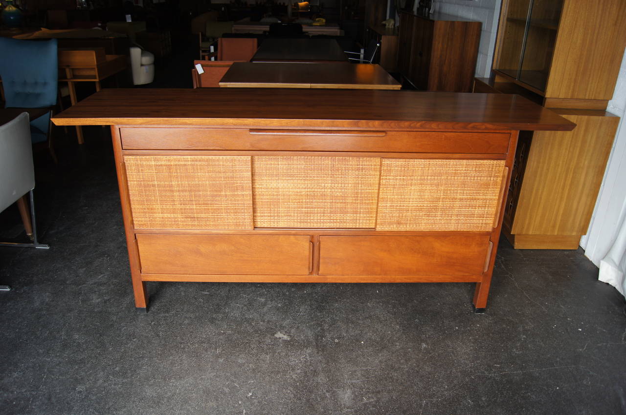 This magnificent sideboard by Edward Wormley comprises an overhanging walnut slab atop a mahogany case with rattan sliding doors and large drawers: one long, shallow drawer and two deeper ones. The three sliding doors open to reveal additional