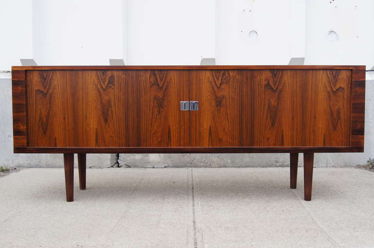 This stately sideboard by Hans Wegner is composed of a rosewood exterior with sliding rosewood tambour doors, an oak interior, and tapered rosewood legs. The interior features numerous drawers and shelves for ample storage.