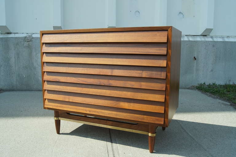 This dresser was designed by Merton Gershun for Martinsville's Dania Collection. It is composed of walnut and with brass detailing along the skirt. It features three drawers with a louvered front and practical dividers. Corresponding tall chest also