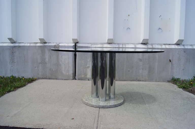 This glass-top dining table features a pedestal base comprised of a granite bottom, four chrome columns, and a chrome top. 