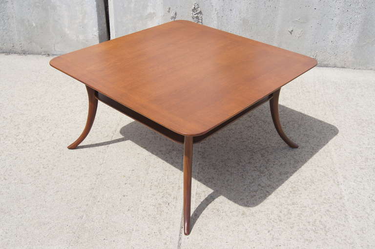 This mahogany coffee table by T.H. Robsjohn-Gibbings for Widdicomb features a rounded square top, a lower shelf, and sweeping saber-shaped legs.
