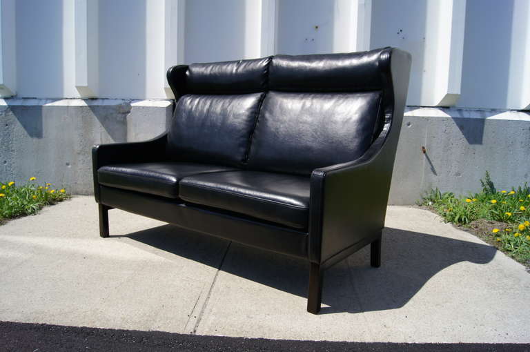 This handsome mid-century Danish modern settee features a curvaceous frame with a comfortable high back and ebonized legs that match the durable black faux leather upholstery — perfect for public spaces or active homes.
