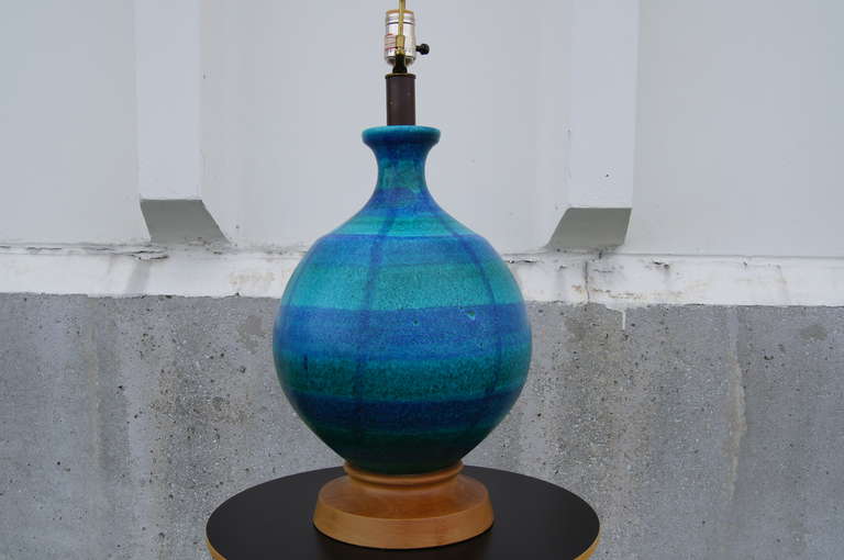 This large Bitossi table lamp comprises a vibrant blue-green spherical ceramic body, a wooden base, and a tall fabric shade with blue ribbon accents. 

The lamp measures 37.5