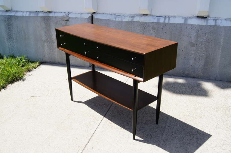 This walnut console table, which features a gently curved front and tapered legs, has an open lower shelf and four drawers with black fronts and small metal drawer pulls.