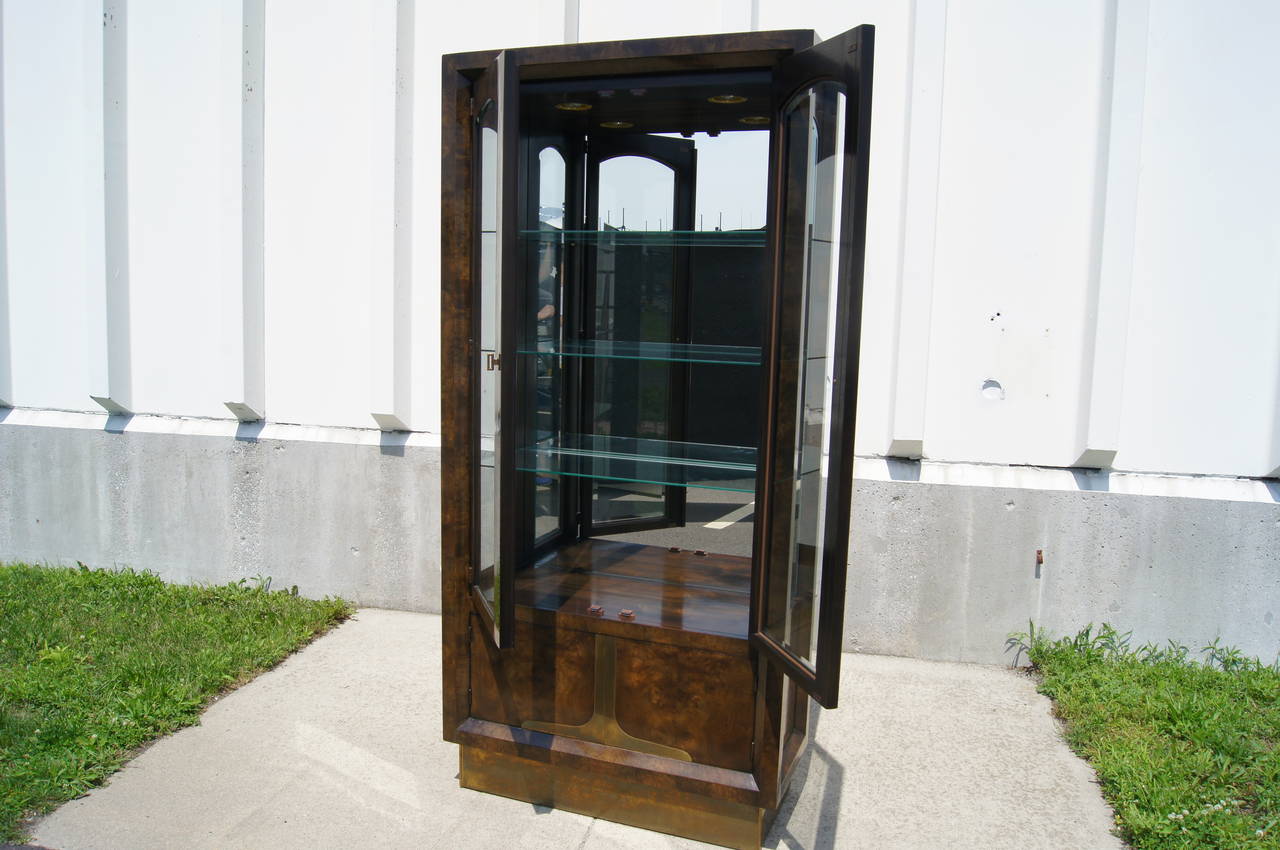 Designed by William Doezema, a cofounder of Mastercraft, this cabinet is composed of dark amboyna burl wood with strong brass accents. It features two glass doors that open to reveal glass shelving, a mirrored back, and two recessed lights for