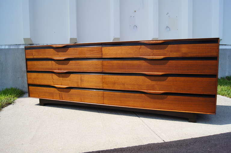 This long walnut dresser was designed by John Kapel and manufactured by John Stuart Inc. It has each drawers, each with a elegant wooden drawer pull, that each feature multiple compartments. 

Corresponding tall chest and pair of nightstands also