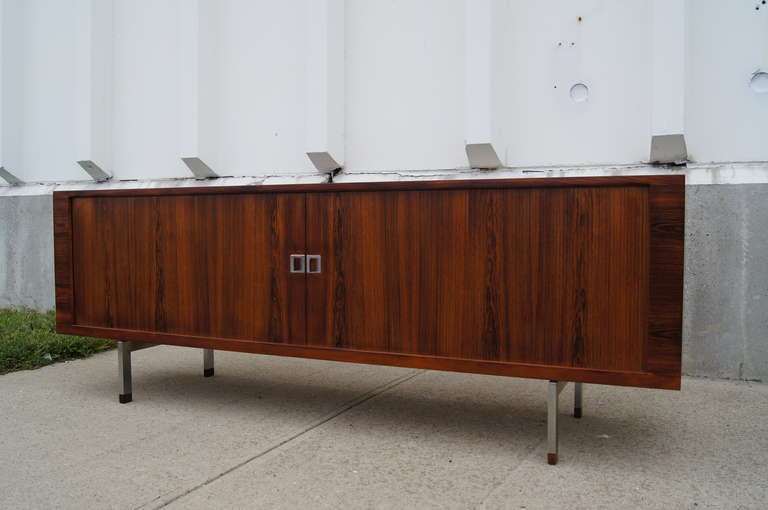 This stately sideboard by Hans Wegner is composed of a rosewood exterior with sliding rosewood tambour doors, an oak interior, and geometric steel legs. The interior features numerous drawers and shelves for ample storage.