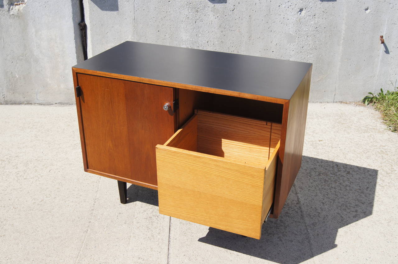 Florence Knoll designed this petit cabinet for Knoll in the 1960s. Perfect for small spaces, it features a walnut case with a black laminate top, ebonized wood legs, leather door pulls, and an oak interior. The lockable sliding doors conceal three