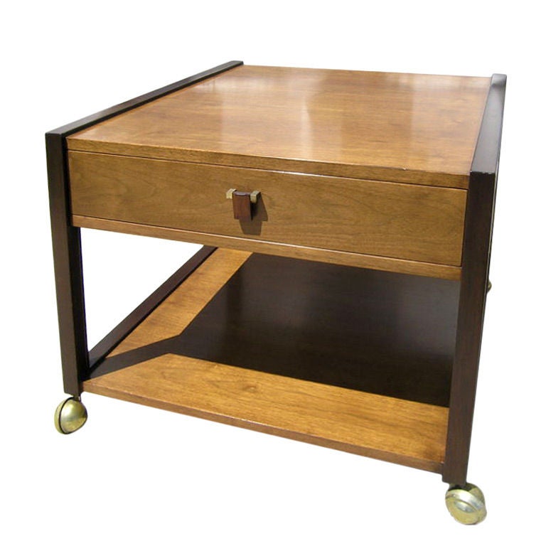 Edward Wormley designed this mahogany side table for Dunbar. The case, whose warm honey tone contrasts with the dark-stained frame, features a wide drawer with a rosewood and brass pull and a deep shelf below. The table sits on bold ball casters in