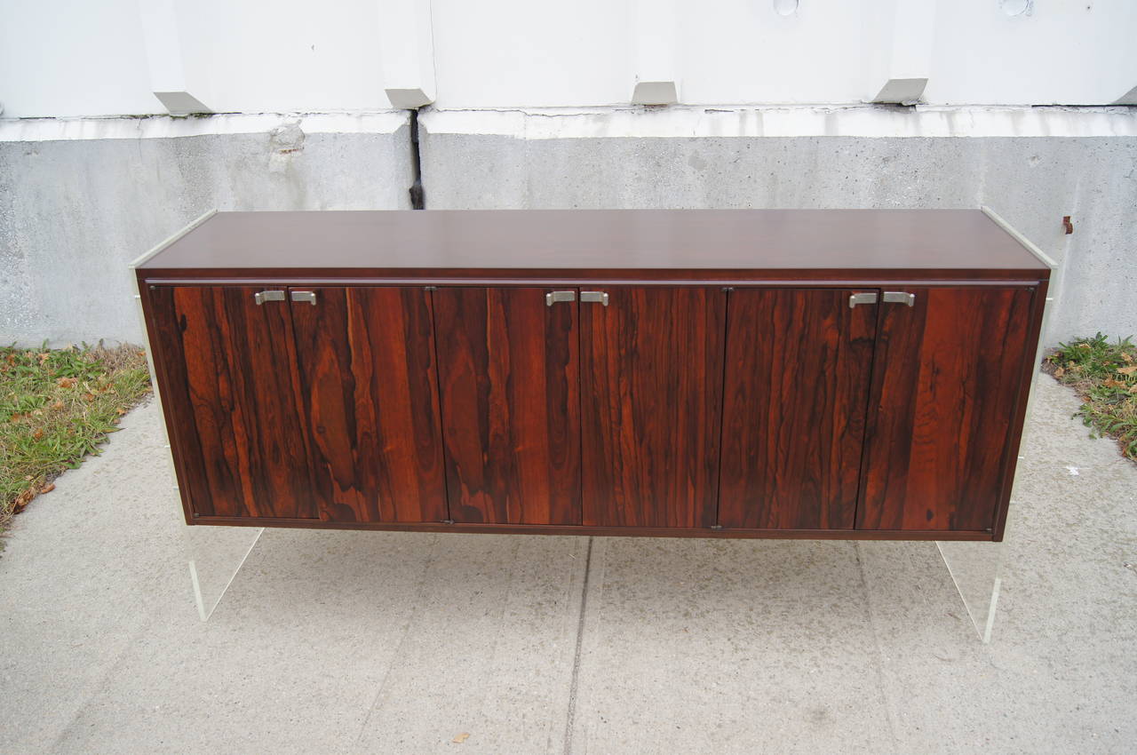 This Milo Baughman sideboard is constructed of a walnut case with rosewood doors and Lucite ends, which give the illusion that the sideboard is floating. It features six doors that house ample storage, including two felt-lined flatware drawers.