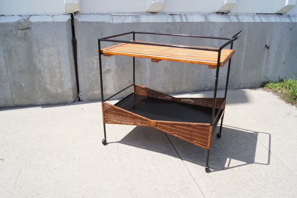 Designed by Arthur Umanoff for Raymor, this tea cart comprises a wrought-iron frame, a slatted wood upper shelf, and a lower shelf in black formica with butterfly basketweave sides.