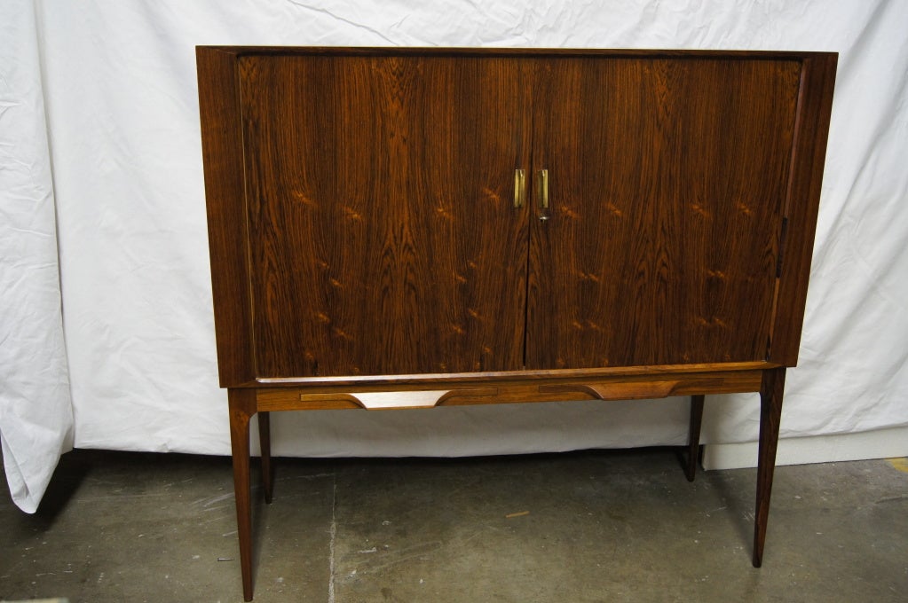This rosewood bar by Illum Wikkelsø has two tambour doors that open to reveal two inner sections. One area has two shallow drawers and two shelves. The other area has one glass shelf and is lined with diamond frosted mirrors and a black glass