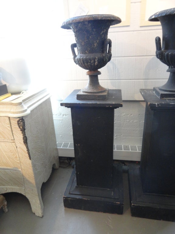 Pair of black iron urns with handles on painted black pedestals. As shown in images, one urn is more recently painted while the other is more antiqued. 

Measures: Urns: 8